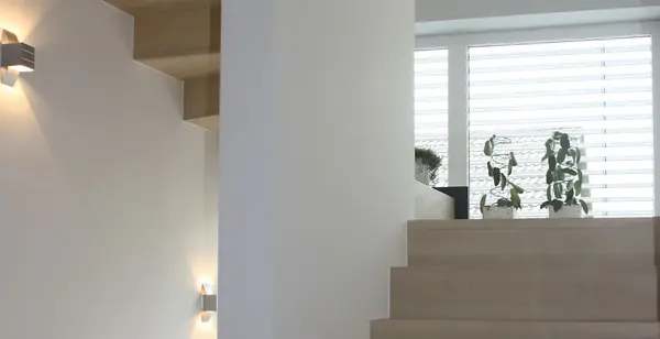 Helle, moderne Holztreppe mit Pflanzen vor großen Fenstern und stilvoller Wandbeleuchtung.